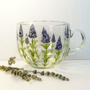 This hand-painted glass mug features a lavender floral design. The sides of the mug are painted with purple lavender flowers and green stems with green leaves and drops of dew. The mug has a curved shape and a clear glass handle. The lavender design is hand-painted and adds a touch of elegance to the mug. This charming mug is perfect for tea, coffee, or hot chocolate.