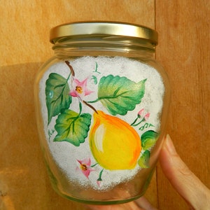 Può includere: Un barattolo di vetro trasparente con coperchio dorato, riempito con contenuto bianco. Il barattolo è dipinto a mano con una pera gialla, foglie verdi e fiori rosa. Il barattolo è tenuto davanti a uno sfondo di legno.