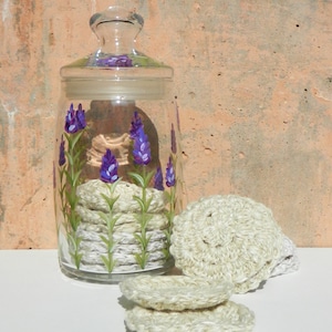 Puede incluir: Un tarro de vidrio con tapa, decorado con flores de lavanda moradas, lleno de rondas de algodón de crochet blancas.