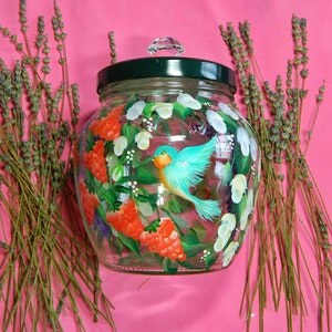 May include: A glass jar with a green lid and a painted design of a blue bird, red berries, and white flowers. The jar is sitting on a pink surface.