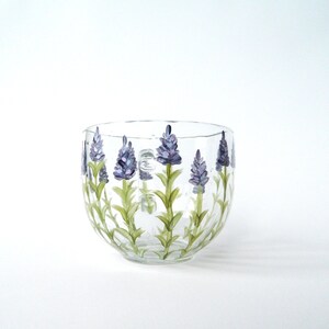 Hand-painted glass mug features a charming design of lavender flowers in shades of purple and green. The flowers are artistically arranged around the mug's body, adding a touch of elegance. The clear glass allows the painted details to stand out beautifully. This lovely mug is perfect for enjoying your favorite beverage and adding a touch of nature to your daily routine. The round shape and clear glass make it a versatile piece for any kitchen or dining setting.