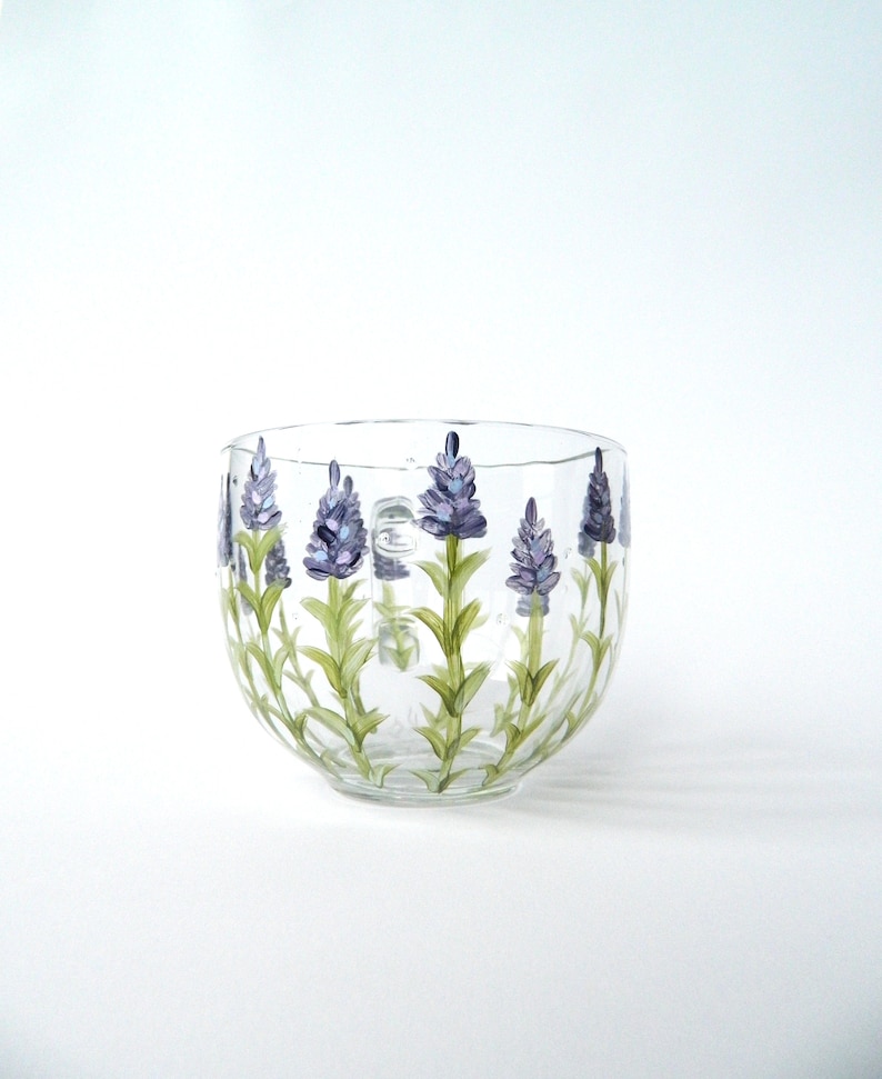 Hand-painted glass mug features a charming design of lavender flowers in shades of purple and green. The flowers are artistically arranged around the mug's body, adding a touch of elegance. The clear glass allows the painted details to stand out beautifully. This lovely mug is perfect for enjoying your favorite beverage and adding a touch of nature to your daily routine. The round shape and clear glass make it a versatile piece for any kitchen or dining setting.