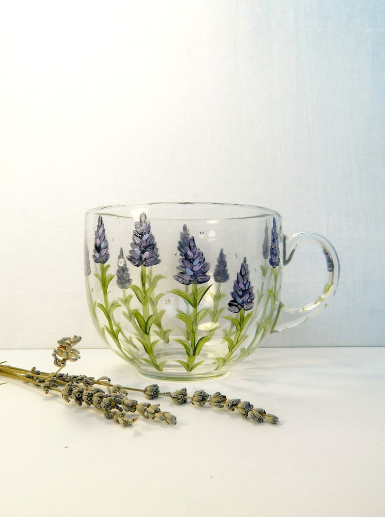 This hand-painted glass mug features a lavender floral design. The sides of the mug are painted with purple lavender flowers and green stems with green leaves and drops of dew. The mug has a curved shape and a clear glass handle. The lavender design is hand-painted and adds a touch of elegance to the mug. This charming mug is perfect for tea, coffee, or hot chocolate.