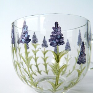 Hand-painted glass mug features a charming lavender design.  Delicate purple lavender flowers and green stems are artfully painted onto the clear glass, creating a lovely botanical theme. The mug's curved shape and transparent nature showcase the painted details beautifully. This hand-painted glass mug is perfect for adding a touch of elegance to any kitchen or tea party.