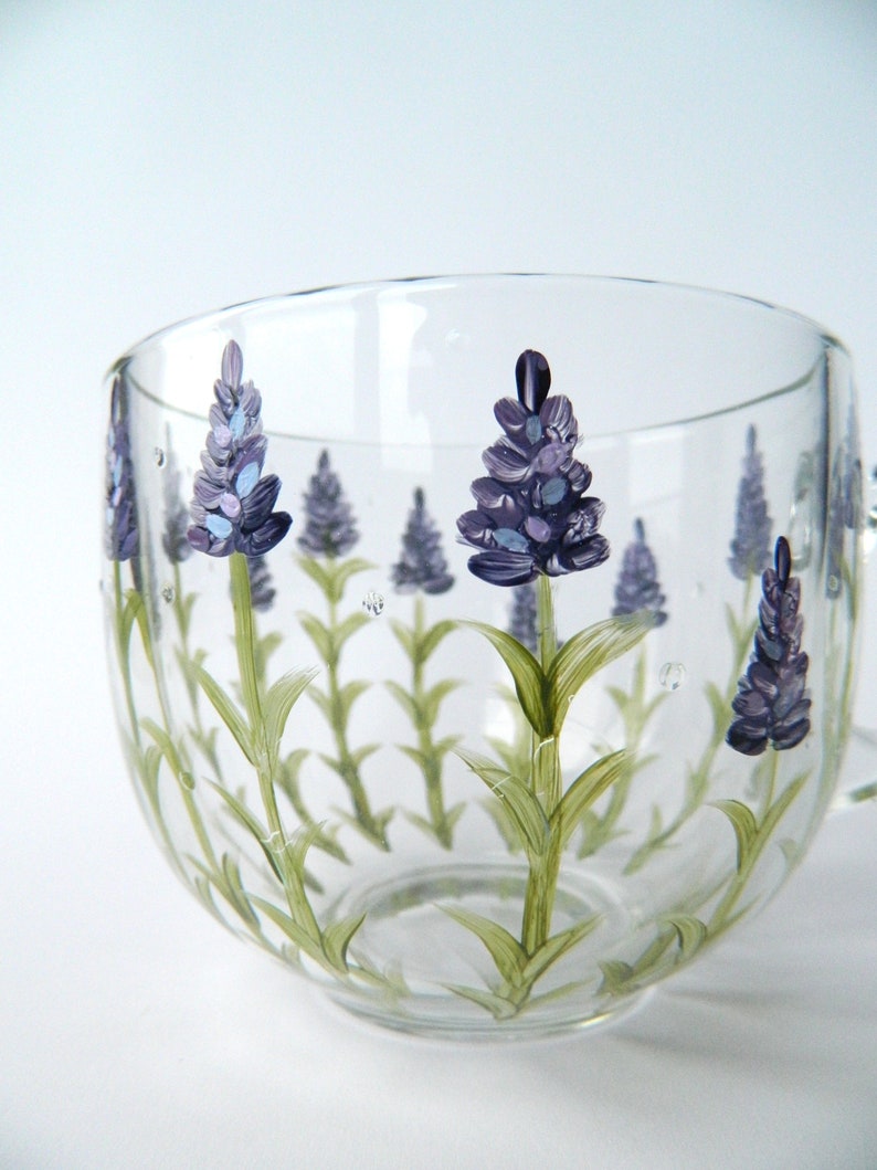 Hand-painted glass mug features a charming lavender design.  Delicate purple lavender flowers and green stems are artfully painted onto the clear glass, creating a lovely botanical theme. The mug's curved shape and transparent nature showcase the painted details beautifully. This hand-painted glass mug is perfect for adding a touch of elegance to any kitchen or tea party.