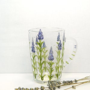 Hand-painted glass mug featuring a lavender floral design. The mug is transparent, showcasing the painted lavender sprigs with purple flowers and green stems against the clear glass.  The mug has a transparent handle, also painted.