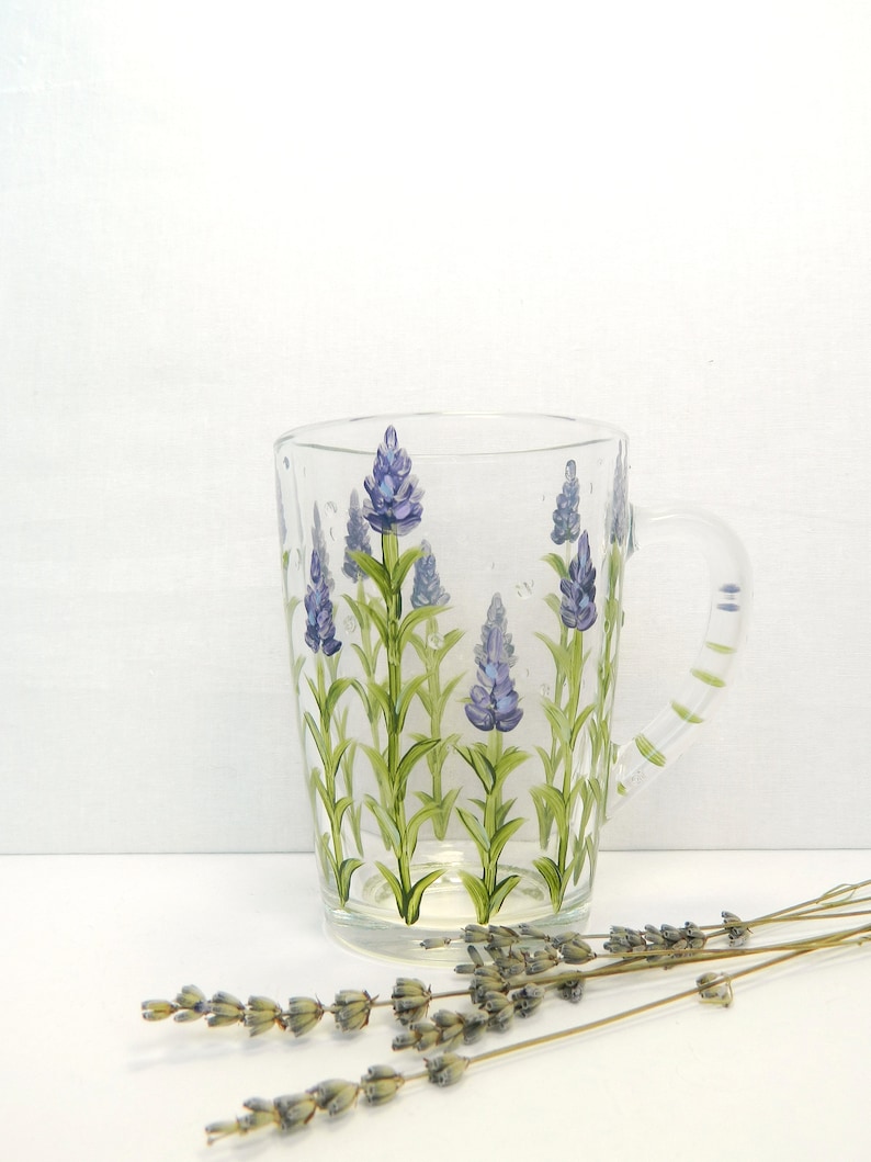 Hand-painted glass mug featuring a lavender floral design. The mug is transparent, showcasing the painted lavender sprigs with purple flowers and green stems against the clear glass.  The mug has a transparent handle, also painted.