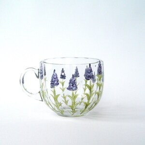A clear glass mug with a handle, decorated with hand-painted lavender flowers. The lavender blossoms are depicted in shades of purple, set against green stems. The mug's design is simple and elegant, suitable for tea or coffee. The glass is transparent, allowing the painted details to be visible. The mug is a charming and functional piece, perfect for adding a touch of nature to any kitchen or dining setting.