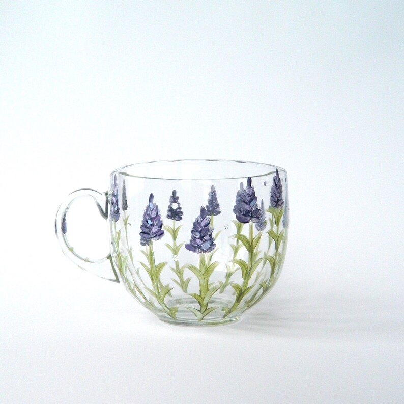 A clear glass mug with a handle, decorated with hand-painted lavender flowers. The lavender blossoms are depicted in shades of purple, set against green stems. The mug's design is simple and elegant, suitable for tea or coffee. The glass is transparent, allowing the painted details to be visible. The mug is a charming and functional piece, perfect for adding a touch of nature to any kitchen or dining setting.