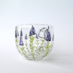 A clear glass mug with a handle features hand-painted lavender flowers. The purple lavender blossoms and green stems encircle the mug, extending onto the handle. The design is simple and elegant, suitable for tea or coffee.
