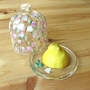 Könnte beinhalten: Eine transparente Glasglocke mit floralem Design, neben einem kleinen Glasteller mit einer Zitrone. Die Glocke ist mit rosa, weißen und grünen Blumen verziert. Die Zitrone ist gelb und liegt auf einem transparenten Glasteller mit bemalten grünen Blättern und weißen Blumen.