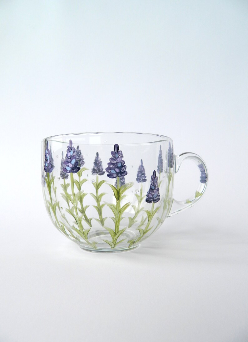 Hand-painted glass mug features a charming lavender design.  The clear glass mug showcases multiple sprigs of lavender, painted in shades of purple and light green. The stems and leaves are delicately rendered, adding a touch of elegance. The mug has a curved shape and a comfortable handle. This lovely mug is perfect for tea, coffee, or hot chocolate, adding a touch of nature to your daily routine.