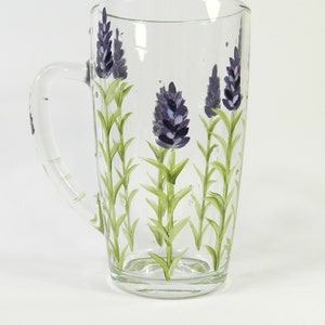 Hand-painted glass mug features a lavender design.  The clear glass mug has a curved handle and showcases multiple sprigs of lavender painted in shades of purple and green. The lavender design is vertically oriented on the mug's surface. This charming mug is perfect for tea, coffee, or other hot beverages.