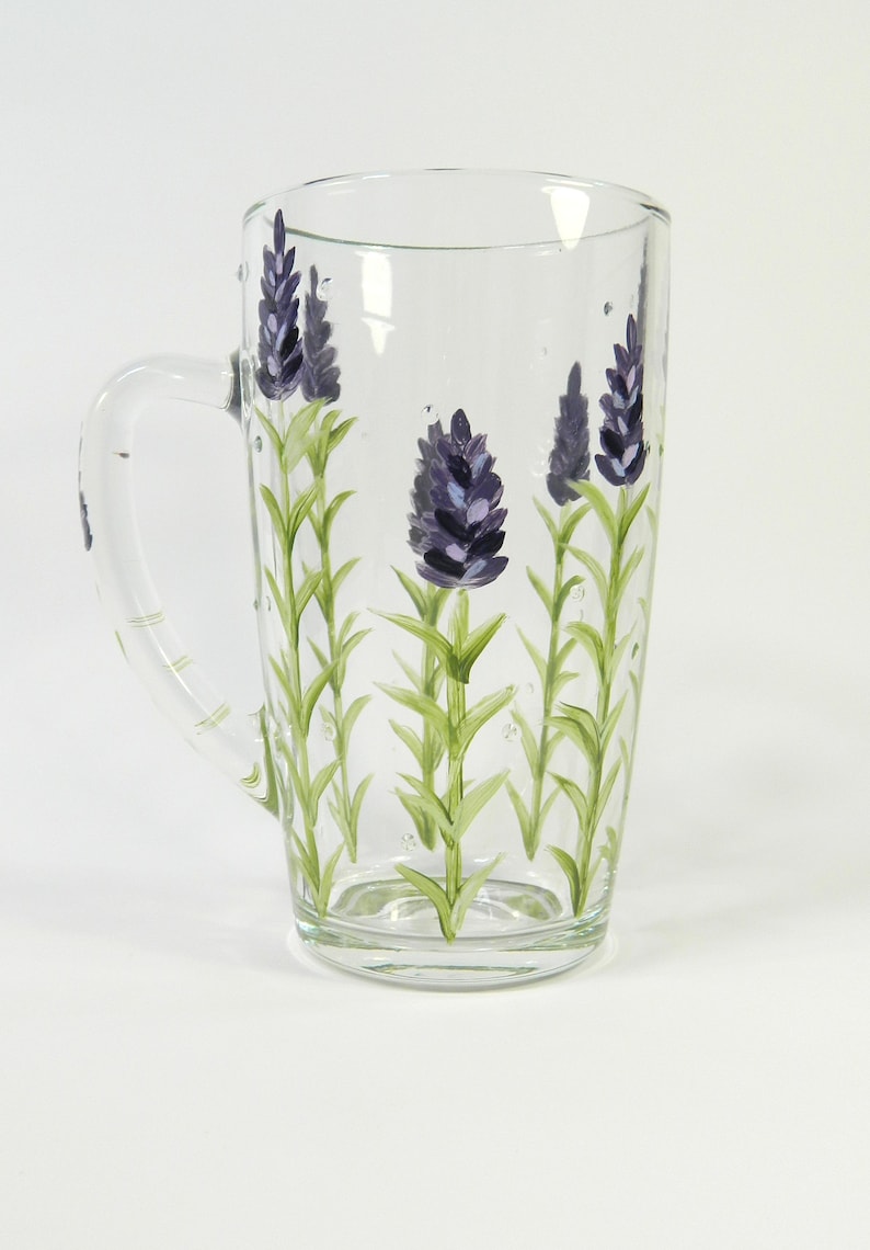 Hand-painted glass mug features a lavender design.  The clear glass mug has a curved handle and showcases multiple sprigs of lavender painted in shades of purple and green. The lavender design is vertically oriented on the mug's surface. This charming mug is perfect for tea, coffee, or other hot beverages.