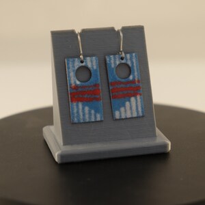 May include: A pair of blue and red enamel earrings with a white pattern and a circle cutout. The earrings are hanging from a silver stand.