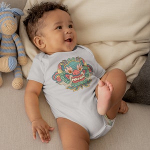 May include: A baby wearing a light blue bodysuit with a colorful graphic print of a mythical creature with a fierce expression. The creature has a green face with red eyes, a yellow mouth with sharp teeth, and a pink nose. The creature is surrounded by swirling clouds and flowers.