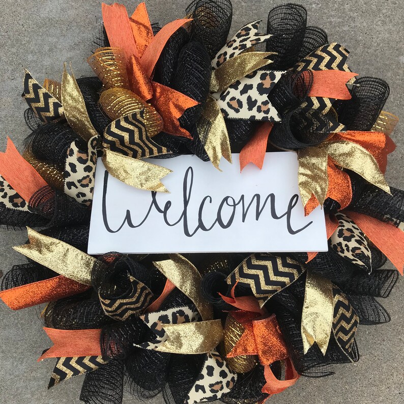 May include: A black, gold, and orange wreath with a welcome sign. The wreath is made of mesh, ribbon, and burlap. It features a leopard print pattern and a chevron pattern.
