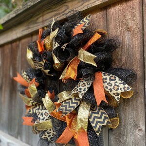 May include: A black, orange, gold, and brown wreath with a leopard print, chevron, and glitter ribbon. The wreath is made of mesh and is hanging on a wooden fence.