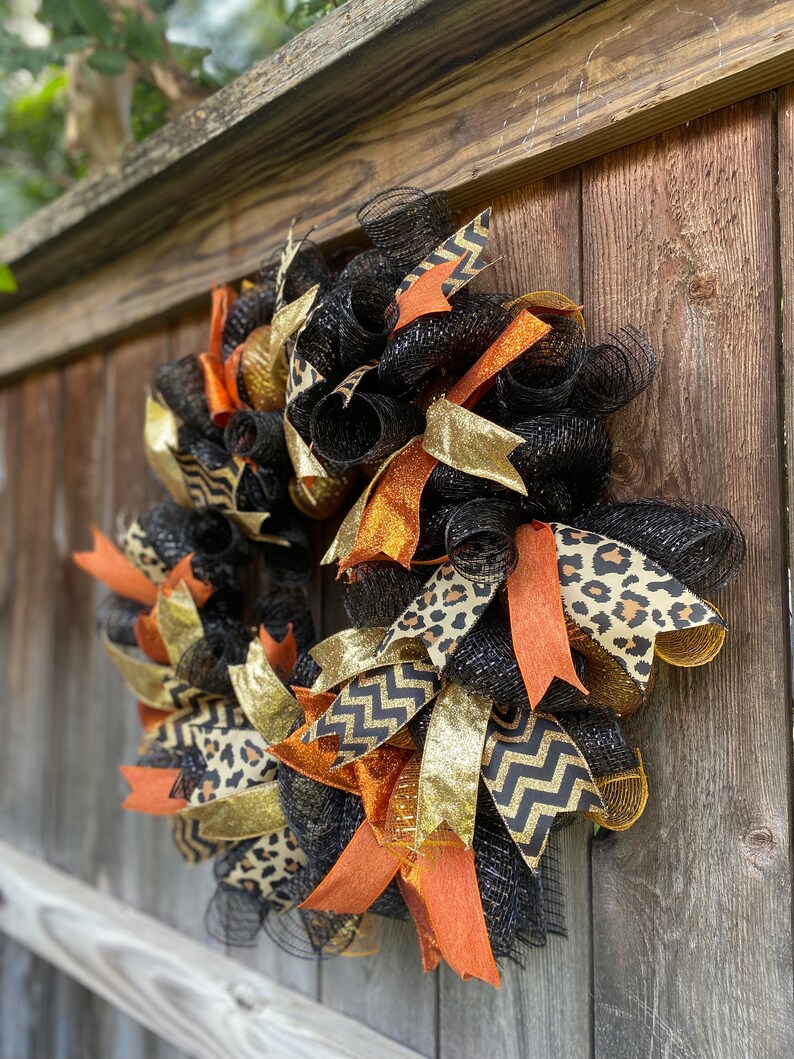 May include: A black, orange, gold, and brown wreath with a leopard print, chevron, and glitter ribbon. The wreath is made of mesh and is hanging on a wooden fence.