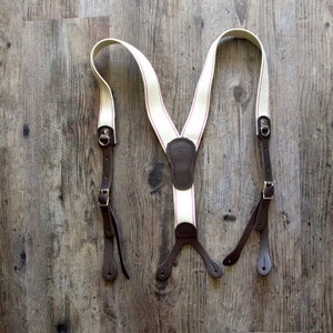 May include: White suspenders with red stripes and brown leather accents. The suspenders have adjustable straps and metal clips.