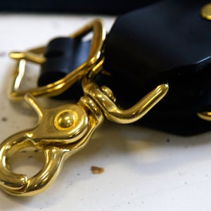 May include: Close-up of a gold-toned metal hook and clasp attached to a black leather strap.