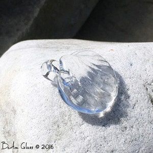 Puede incluir: Un colgante de concha de mar de vidrio transparente con una superficie texturizada. El colgante está apoyado en una roca blanca.