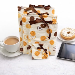 May include: A set of beige fabric bags with a bagel pattern, tied with brown ribbon. A cup of coffee, a bagel with cream cheese, and a tablet are also present, suggesting a breakfast theme.