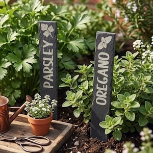 May include: Two black slate garden markers with white text reading "PARSLEY" and "OREGANO". The markers are in a garden bed with green herbs and a small terracotta pot with white flowers. A wooden tray with gardening tools is in the foreground.
