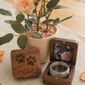 May include: A wooden pet memorial box with a clear glass jar inside and a photo of a dog. The box is open, revealing the jar and photo. A separate wooden square with paw prints and the name "CHARLIE" is next to the box. A potted plant with pink flowers is in the background.