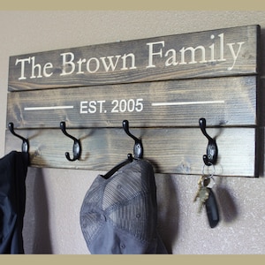 May include: A wooden wall-mounted coat rack with the text "The Brown Family" and "EST. 2005" in white. It has five black metal hooks, with a jacket, a hat, and keys hanging from them. The wood has a gray stain.