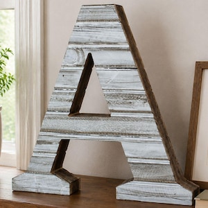 May include: A large wooden letter "A" with a distressed, whitewashed finish. The letter is constructed from horizontal planks, with a natural wood-toned edge. It stands on a wooden surface, near a window and a framed picture.