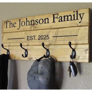 May include: A wooden wall-mounted coat rack with four black metal hooks. The sign reads "The Johnson Family EST. 2025" in black lettering. A black jacket, a gray cap, and keys hang from the hooks.