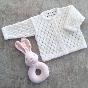 May include: A white long-sleeved knit cardigan featuring an openwork diamond pattern and a round neckline, paired with a soft pink plush rabbit rattle toy with polka-dot fabric on the inner ears and embroidered facial features.