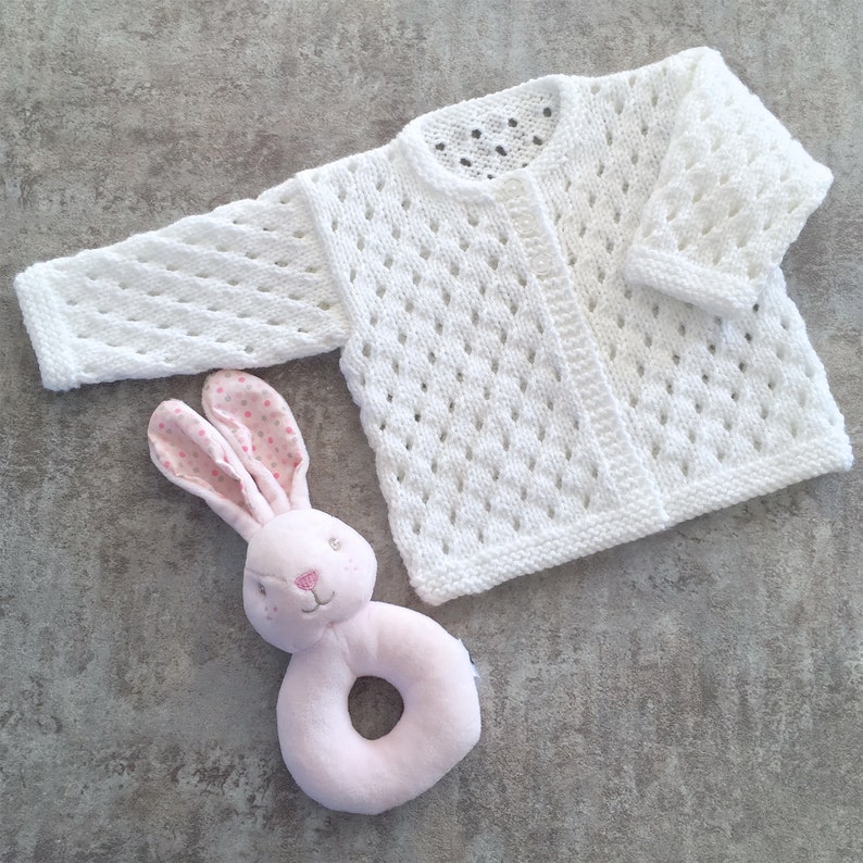 May include: A white long-sleeved knit cardigan featuring an openwork diamond pattern and a round neckline, paired with a soft pink plush rabbit rattle toy with polka-dot fabric on the inner ears and embroidered facial features.