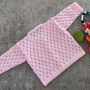 May include: A light pink long-sleeve sweater featuring an openwork diamond knit pattern with a garter stitch border at the cuffs and hem. Beside the sweater is a multi-colored baby rattle toy with a small plush bear head and plastic rings.