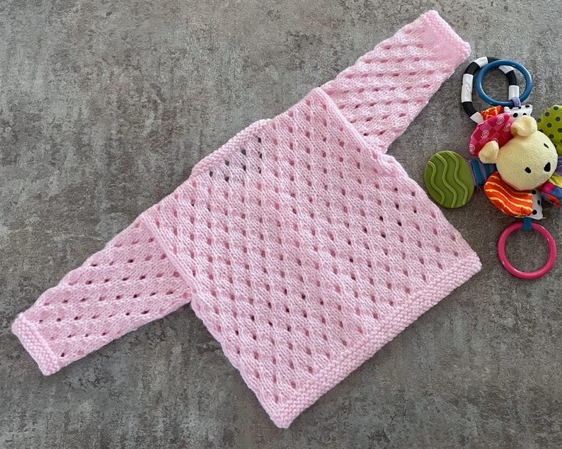 May include: A light pink long-sleeve sweater featuring an openwork diamond knit pattern with a garter stitch border at the cuffs and hem. Beside the sweater is a multi-colored baby rattle toy with a small plush bear head and plastic rings.