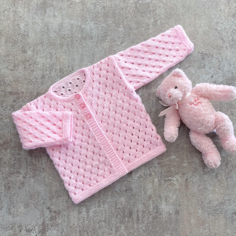 May include: A light pink long-sleeve knit cardigan featuring a diamond-shaped openwork pattern, a round neckline, and a front button closure, accompanied by a small, fuzzy light pink teddy bear with a ribbon tied around its neck.