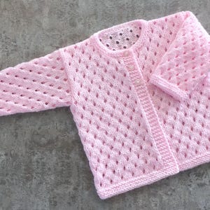 May include: A light pink long-sleeve cardigan featuring an openwork diamond knit pattern throughout the body and sleeves. The garment has a round neckline, a ribbed hem, and a front button closure with small, round, white buttons.