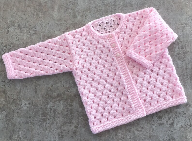 May include: A light pink long-sleeve cardigan featuring an openwork diamond knit pattern throughout the body and sleeves. The garment has a round neckline, a ribbed hem, and a front button closure with small, round, white buttons.