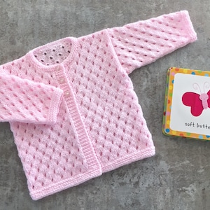 May include: A light pink long-sleeve knit cardigan featuring a delicate diamond-shaped openwork pattern, a round neckline, and two small buttons. Beside it lies a small square book with a pink butterfly illustration and the text soft butterfly.