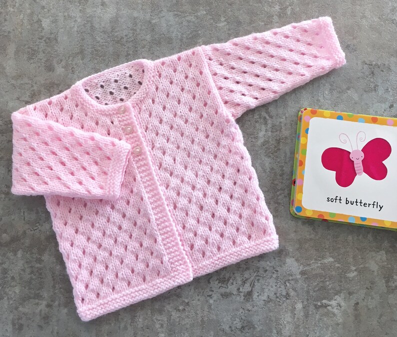 May include: A light pink long-sleeve knit cardigan featuring a delicate diamond-shaped openwork pattern, a round neckline, and two small buttons. Beside it lies a small square book with a pink butterfly illustration and the text soft butterfly.