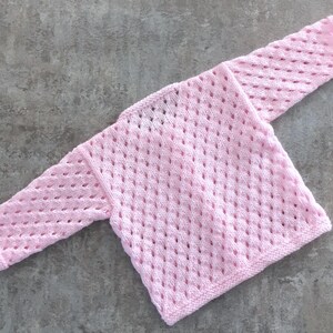 May include: A light pink long-sleeve sweater featuring an openwork diamond pattern throughout the knit fabric. The garment has a simple crew neckline and ribbed cuffs at the ends of the sleeves, showcasing a consistent texture and soft color.