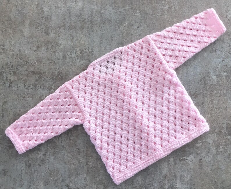 May include: A light pink long-sleeve sweater featuring an openwork diamond pattern throughout the knit fabric. The garment has a simple crew neckline and ribbed cuffs at the ends of the sleeves, showcasing a consistent texture and soft color.