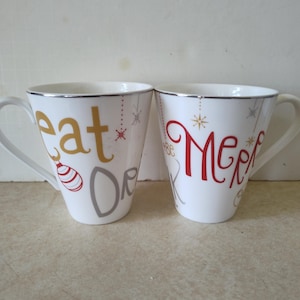 May include: Two white ceramic mugs with silver rims. One mug has the words "eat or" in gold and gray, with a red and white ornament design. The other mug says "Merry" in red with star accents.