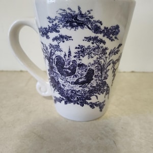 May include: White ceramic mug with a blue toile design featuring a rooster, trees, and other elements. The mug has a curved handle and a slightly flared shape. The design is detailed and covers most of the mug's surface.