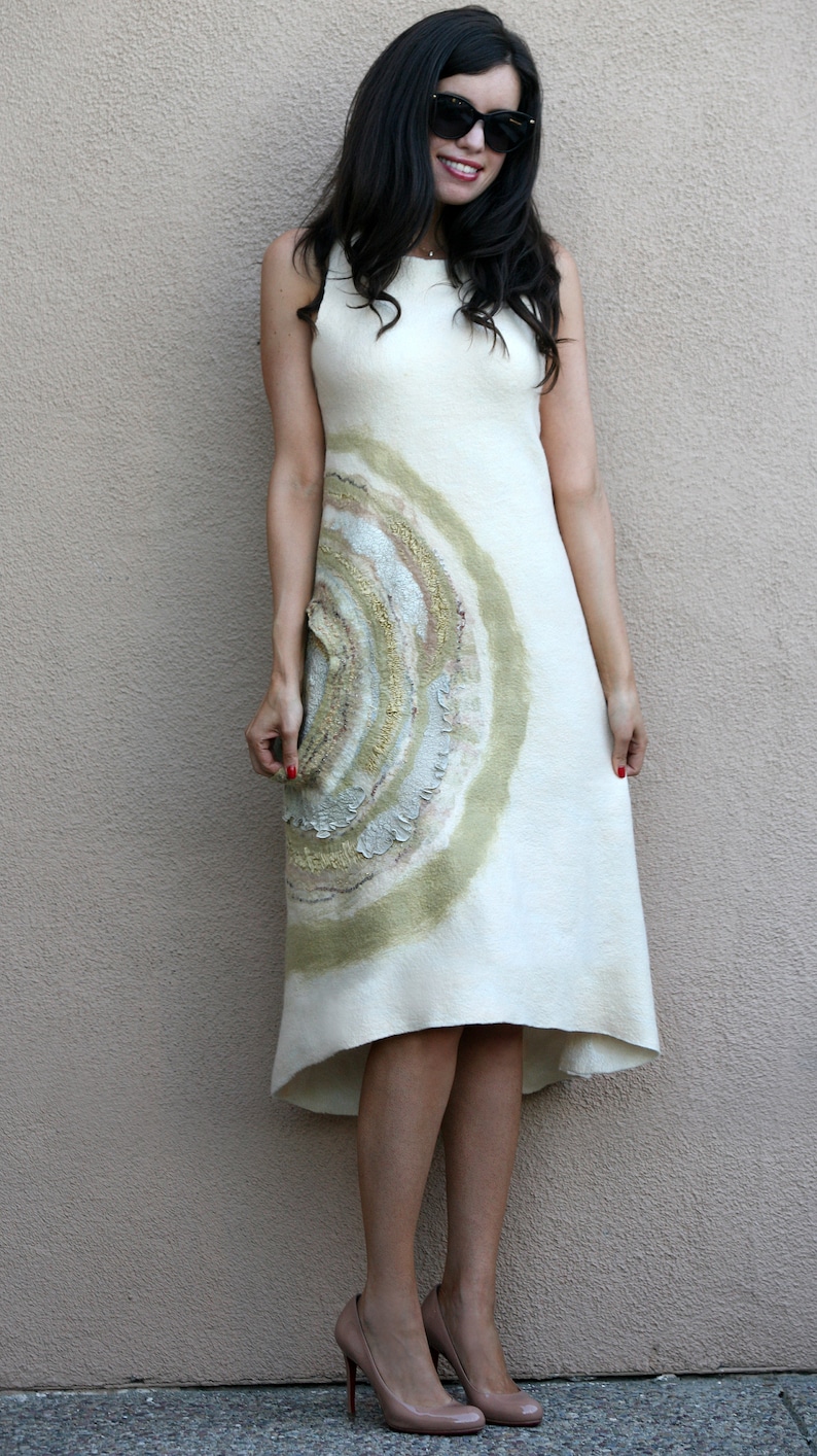 Puede incluir: Una mujer con un vestido blanco con un dise&ntilde;o circular verde y marr&oacute;n. Lleva gafas de sol y tiene el pelo recogido hacia atr&aacute;s. Est&aacute; de pie frente a una pared de color marr&oacute;n claro.