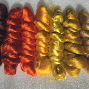 May include: Seven bundles of lustrous fiber arranged in a gradient color sequence. The colors transition from a deep dark brown on the left, through shades of vibrant red, bright orange, and sunny yellow, to a muted golden brown on the right.