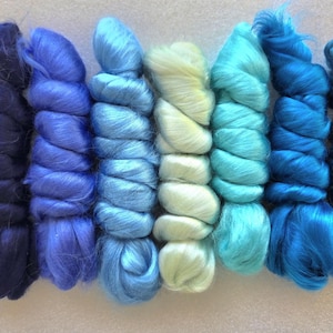 May include: Seven bundles of combed fiber arranged in a row, displaying a gradient of colors from deep navy blue and royal blue to light sky blue, pale yellow-green, bright turquoise, vibrant azure, and a medium teal blue shade.
