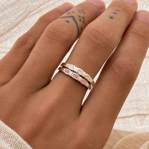 May include: Two stacked metal rings featuring a series of small, rectangular inlays. The top ring contains white, opaque rectangular segments, while the bottom ring features light pink, opaque rectangular segments set within a metallic band.