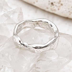May include: A silver ring with an irregular, textured surface and a wavy, organic band shape. The metal has a polished, reflective finish that catches the light, highlighting the uneven, hand-crafted appearance of the circular band structure.
